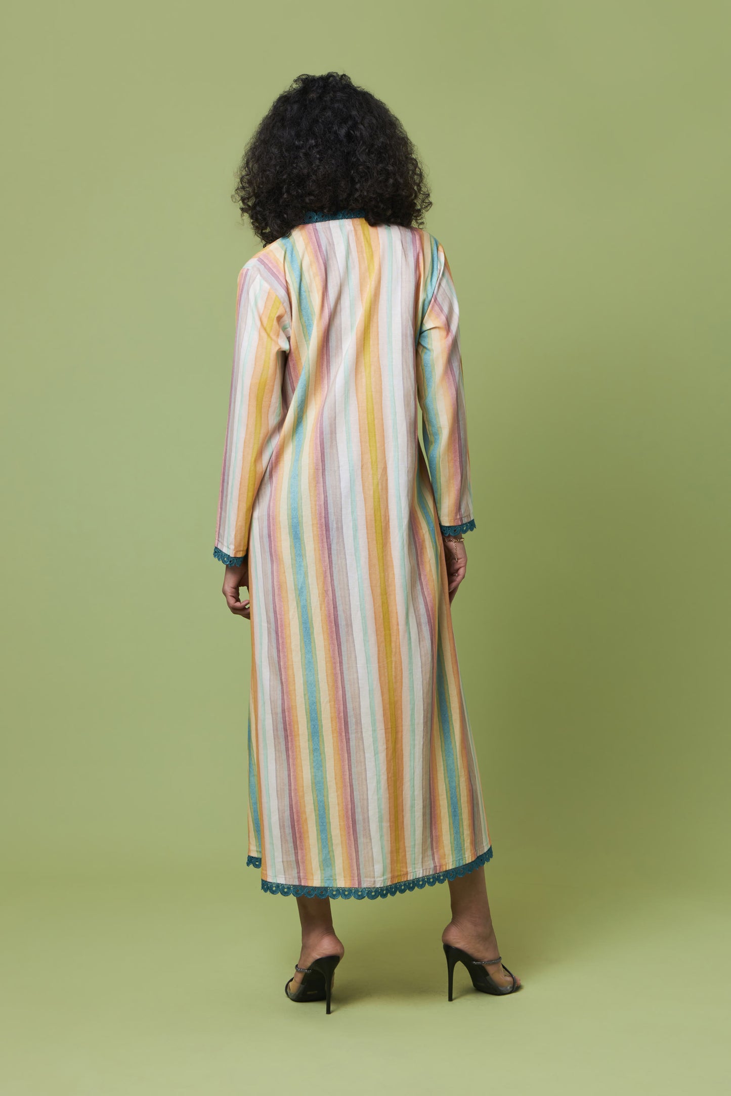 A woman viewed from behind wearing a long-sleeved maxi dress with vertical pastel stripes in yellow, orange, pink, blue, and white, paired with black heels, set against a solid light green studio background.