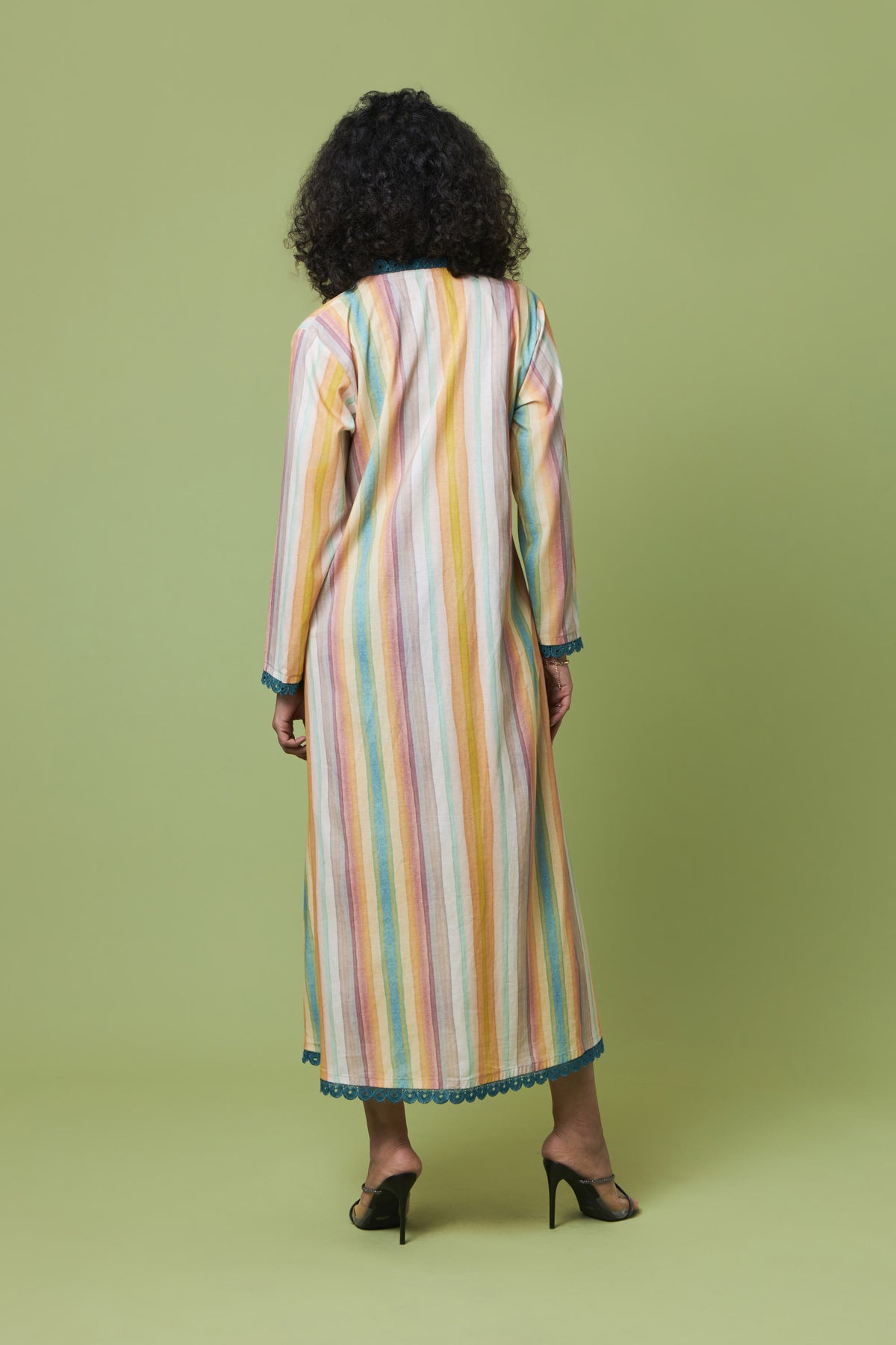 A woman viewed from behind wearing a long-sleeved maxi dress with vertical pastel stripes in yellow, orange, pink, blue, and white, paired with black heels, set against a solid light green studio background.