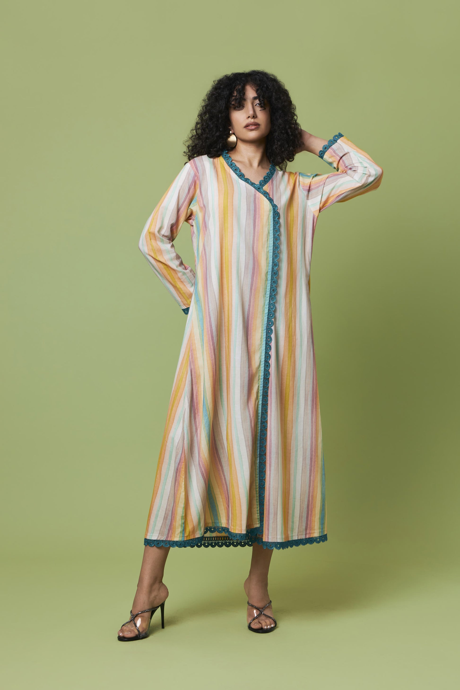 A woman modeling a long-sleeved, midi-length kaftan dress with vertical stripes in yellow, orange, pink, and white, accented with teal trim, set against a solid light green studio background.