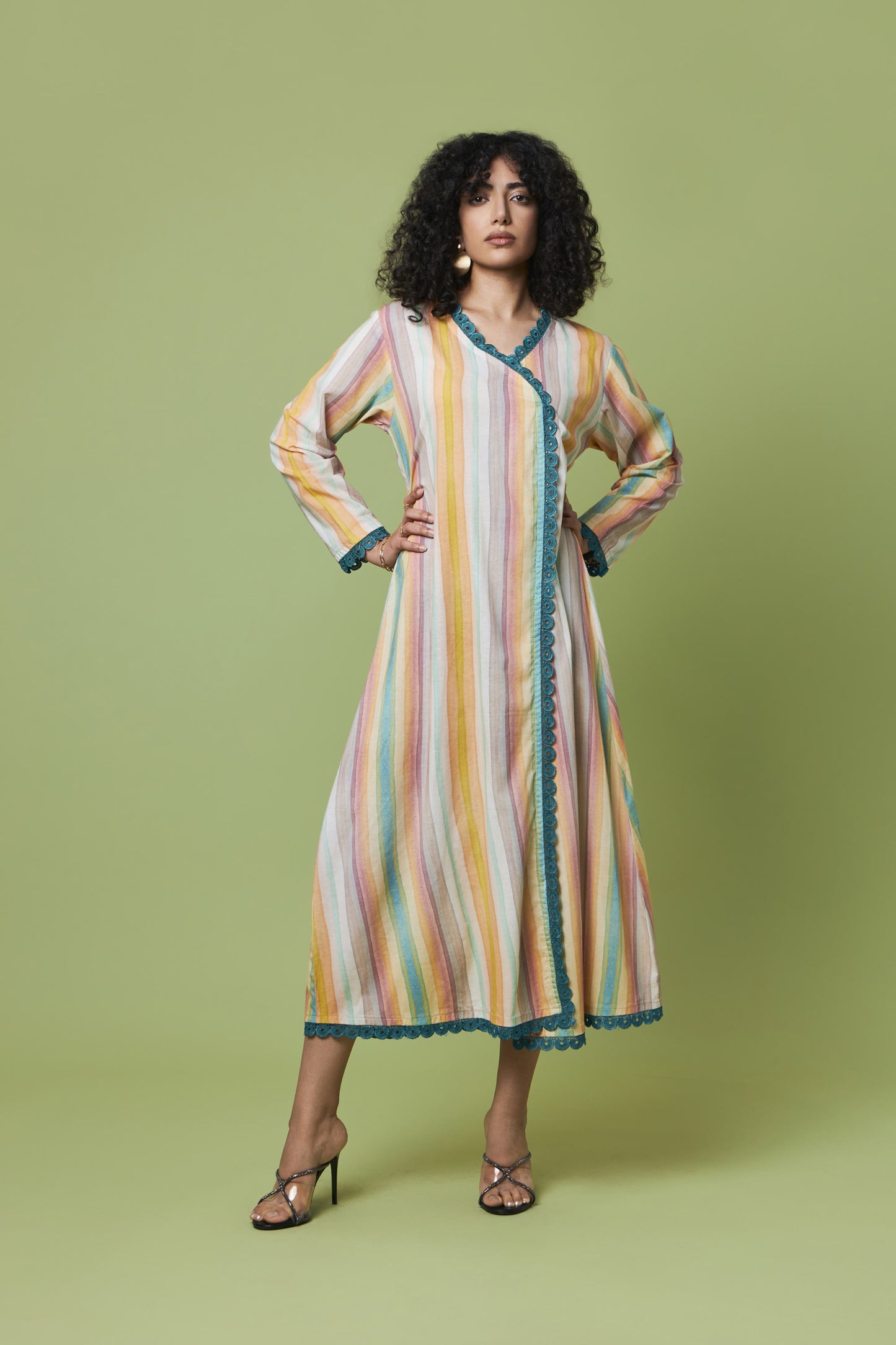 A woman modeling a long-sleeved, wrap-style midi dress with vertical pastel stripes in yellow, orange, pink, and blue, accented with teal lace trim, set against a solid sage green studio background.