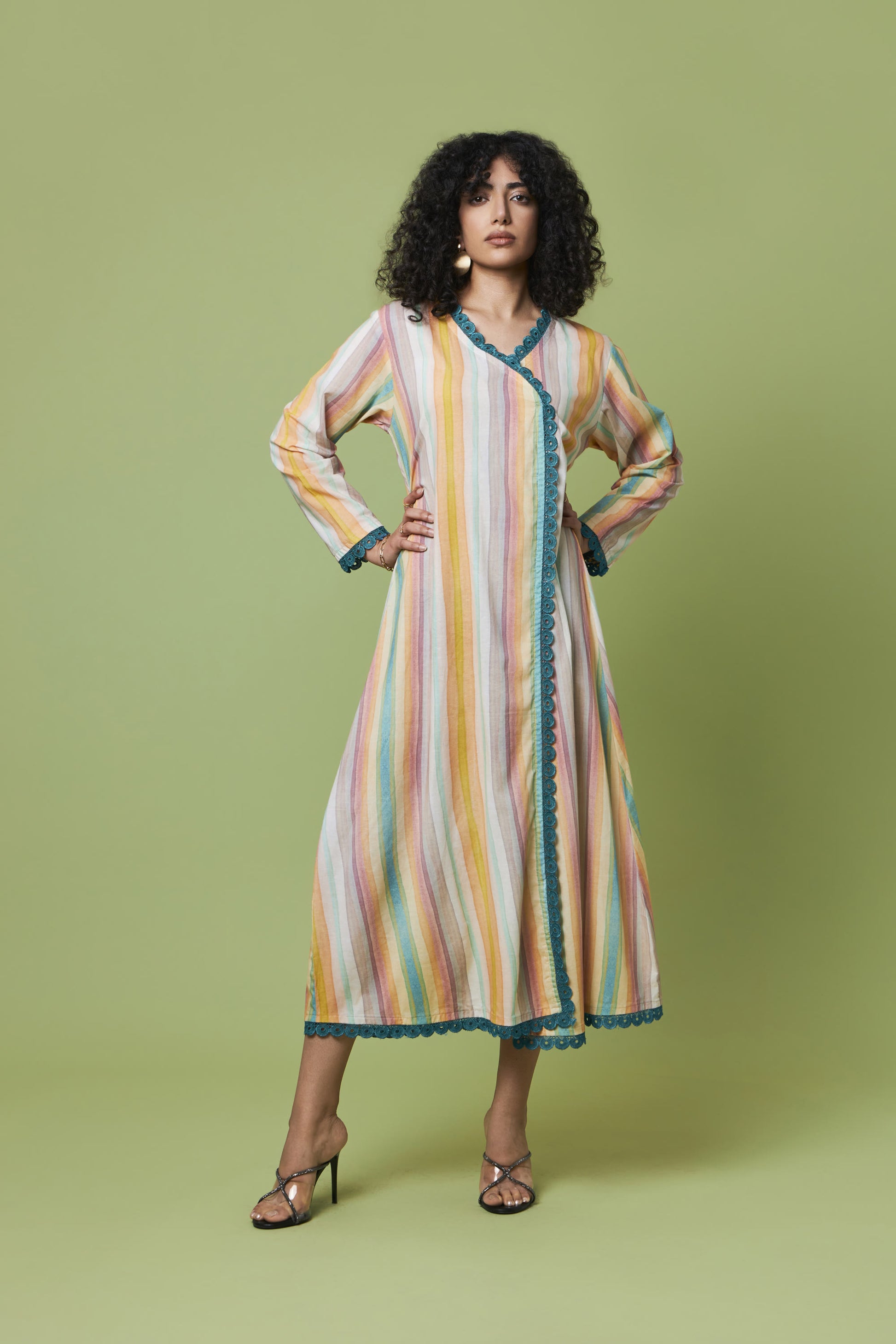 A woman modeling a long-sleeved, wrap-style midi dress with vertical pastel stripes in yellow, orange, pink, and blue, accented with teal lace trim, set against a solid sage green studio background.