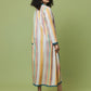 A woman stands in profile wearing a long, loose maxi dress with vertical pastel stripes in yellow, orange, pink, blue, and white. The dress has a high neckline and is styled with black high heels against a solid sage green studio background.