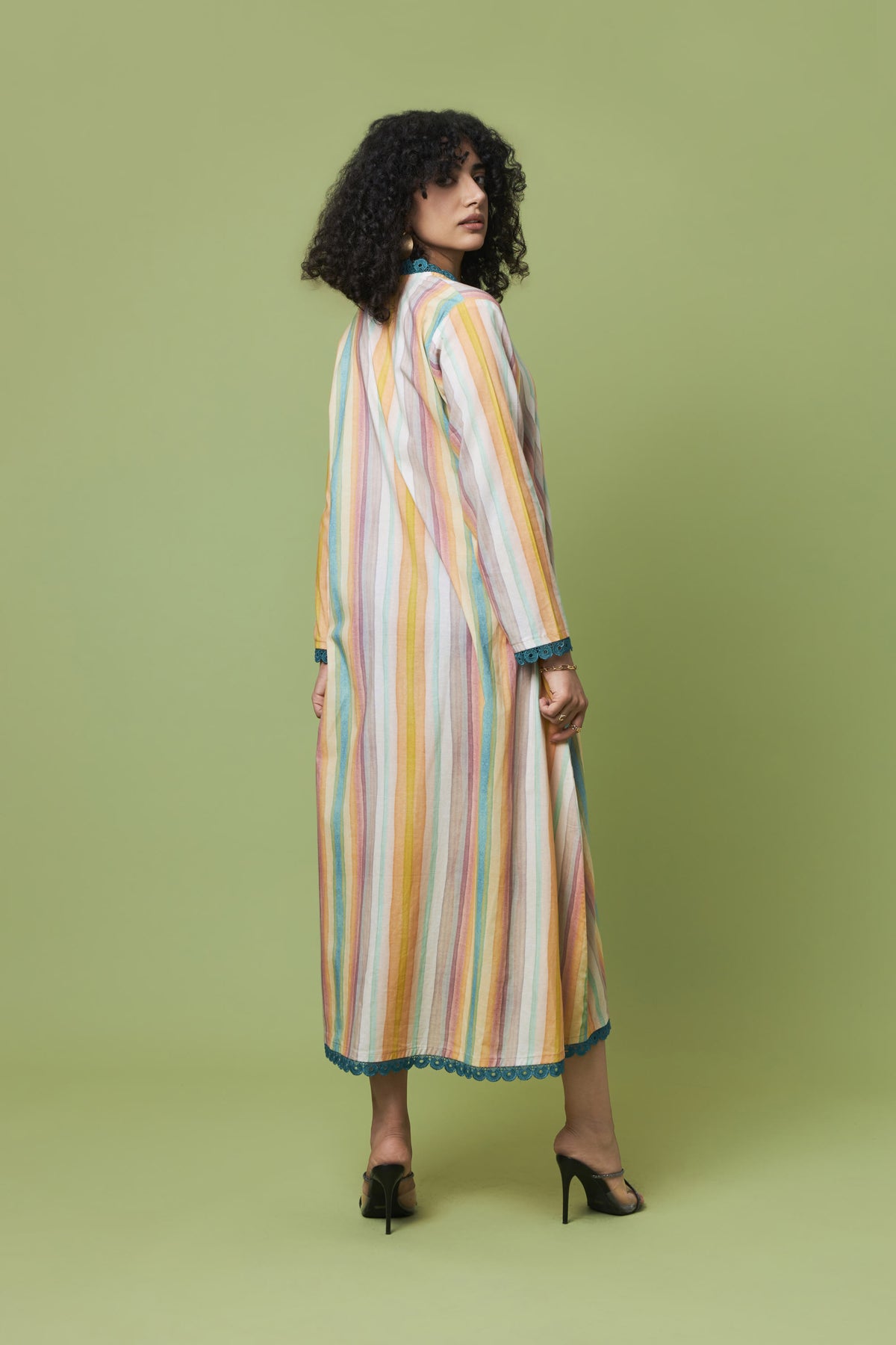 A woman stands in profile wearing a long, loose maxi dress with vertical pastel stripes in yellow, orange, pink, blue, and white. The dress has a high neckline and is styled with black high heels against a solid sage green studio background.