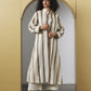 A model with curly black hair stands in a gold-framed arched doorway wearing a long-sleeved, button-up striped tunic in cream and brown, paired with cream cropped pants and brown strappy heels.