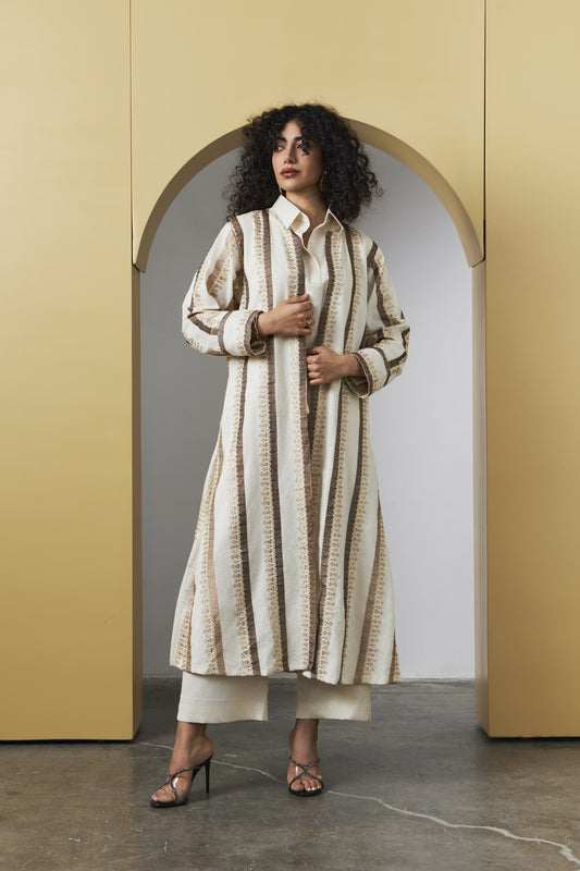 A model with curly black hair stands in a gold-framed arched doorway wearing a long-sleeved, button-up striped tunic in cream and brown, paired with cream cropped pants and brown strappy heels.