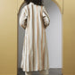 A model viewed from behind wearing a long cream and beige vertically striped coat with textured vertical lines, standing in front of a yellow archway.