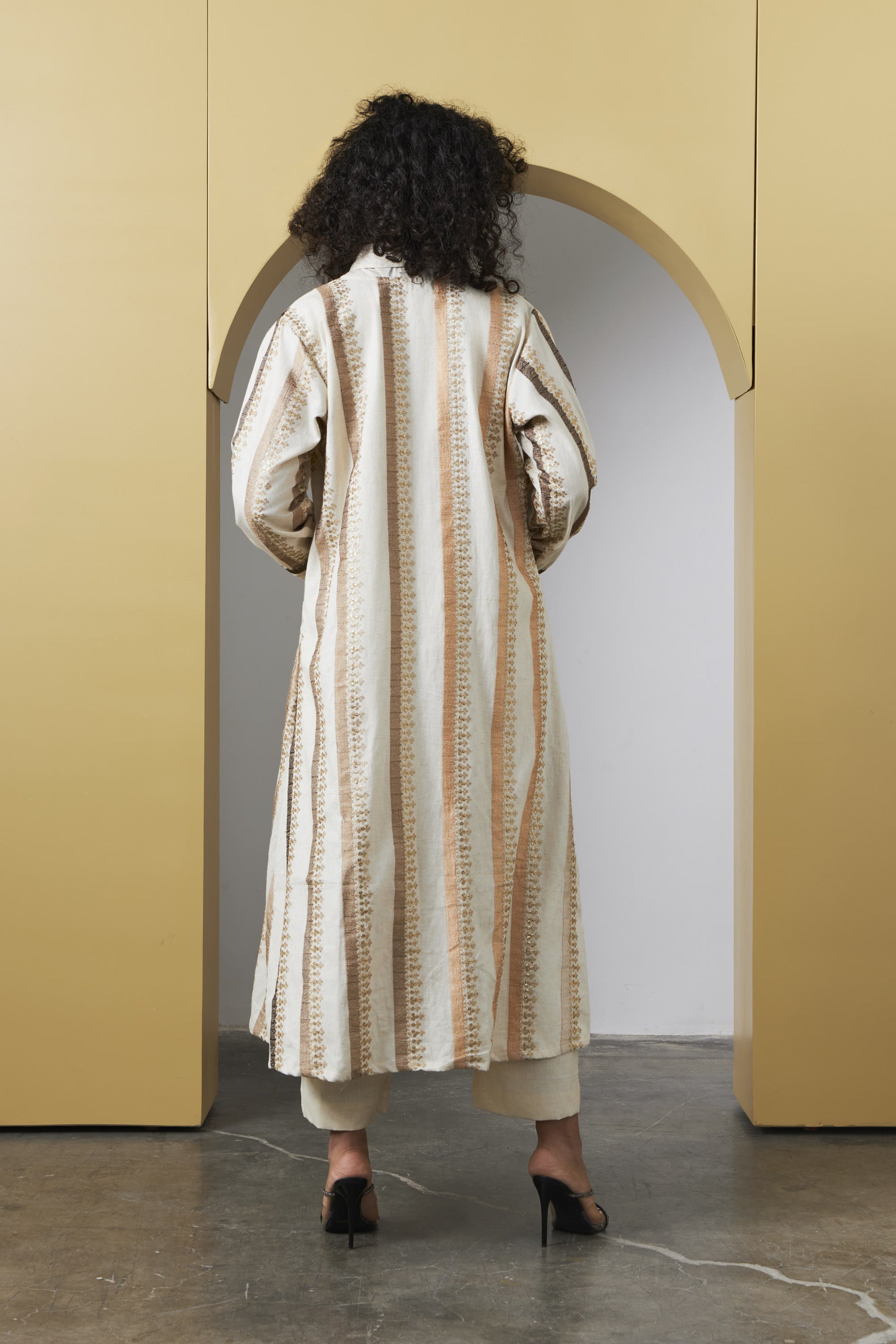 A model viewed from behind wearing a long cream and beige vertically striped coat with textured vertical lines, standing in front of a yellow archway.