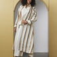 A model with curly dark hair stands in front of a beige arched wall opening, wearing a long cream shirt and matching wide-leg trousers with a long striped cardigan featuring vertical brown and beige textured stripes.