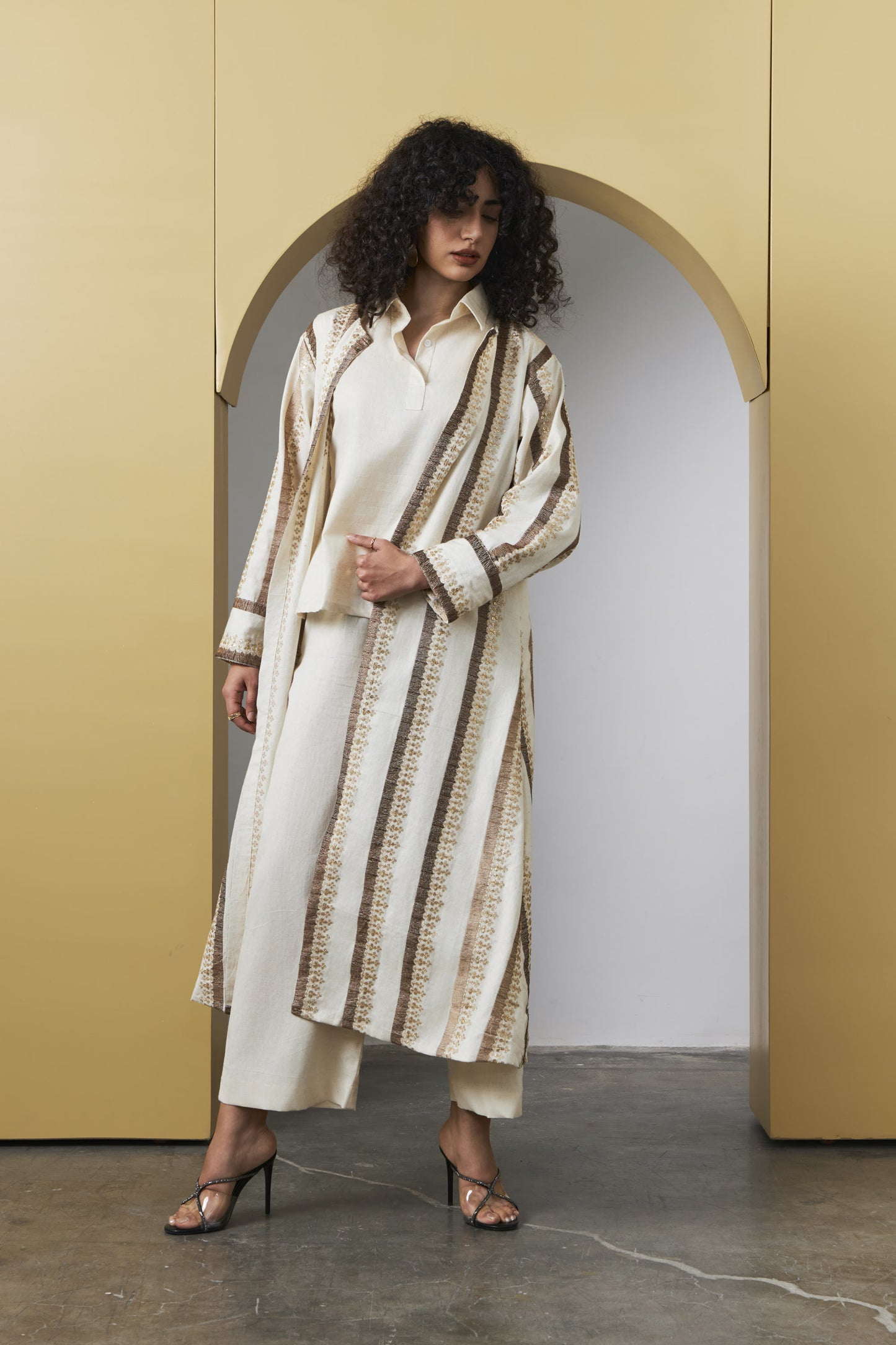 A model with curly dark hair stands in front of a beige arched wall opening, wearing a long cream shirt and matching wide-leg trousers with a long striped cardigan featuring vertical brown and beige textured stripes.