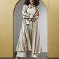 A model stands in front of a yellow arched backdrop wearing a long cream and beige patterned cardigan over a white top and white cropped pants, paired with brown heeled sandals.