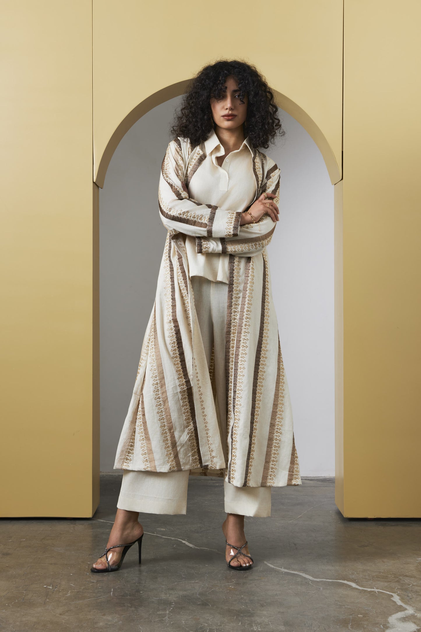 A model stands in front of a yellow arched backdrop wearing a long cream and beige patterned cardigan over a white top and white cropped pants, paired with brown heeled sandals.