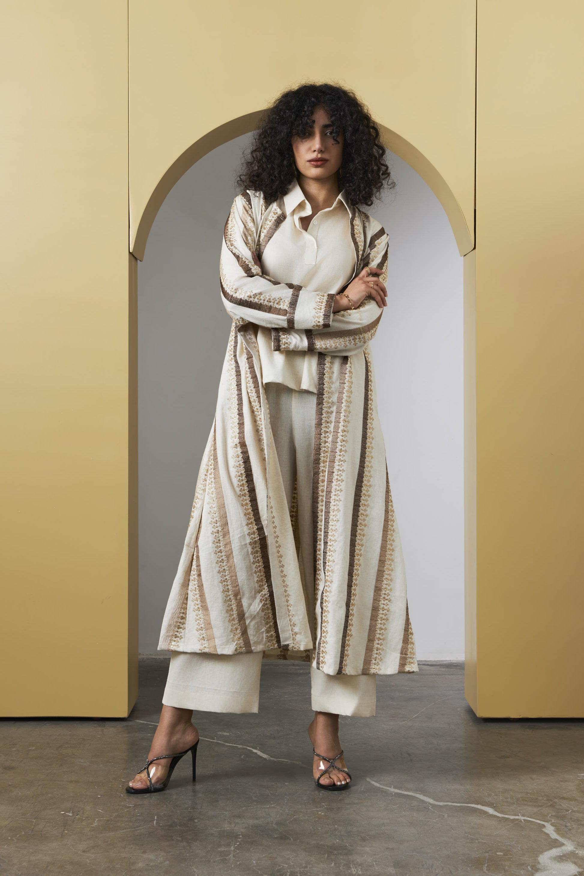 A model stands in front of a yellow arched backdrop wearing a long cream and beige patterned cardigan over a white top and white cropped pants, paired with brown heeled sandals.
