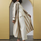A model with curly dark hair stands in a minimalist studio setting with a yellow archway background, wearing a cream and beige patterned long cardigan over a matching cream top and cropped trousers, paired with black strappy heels.