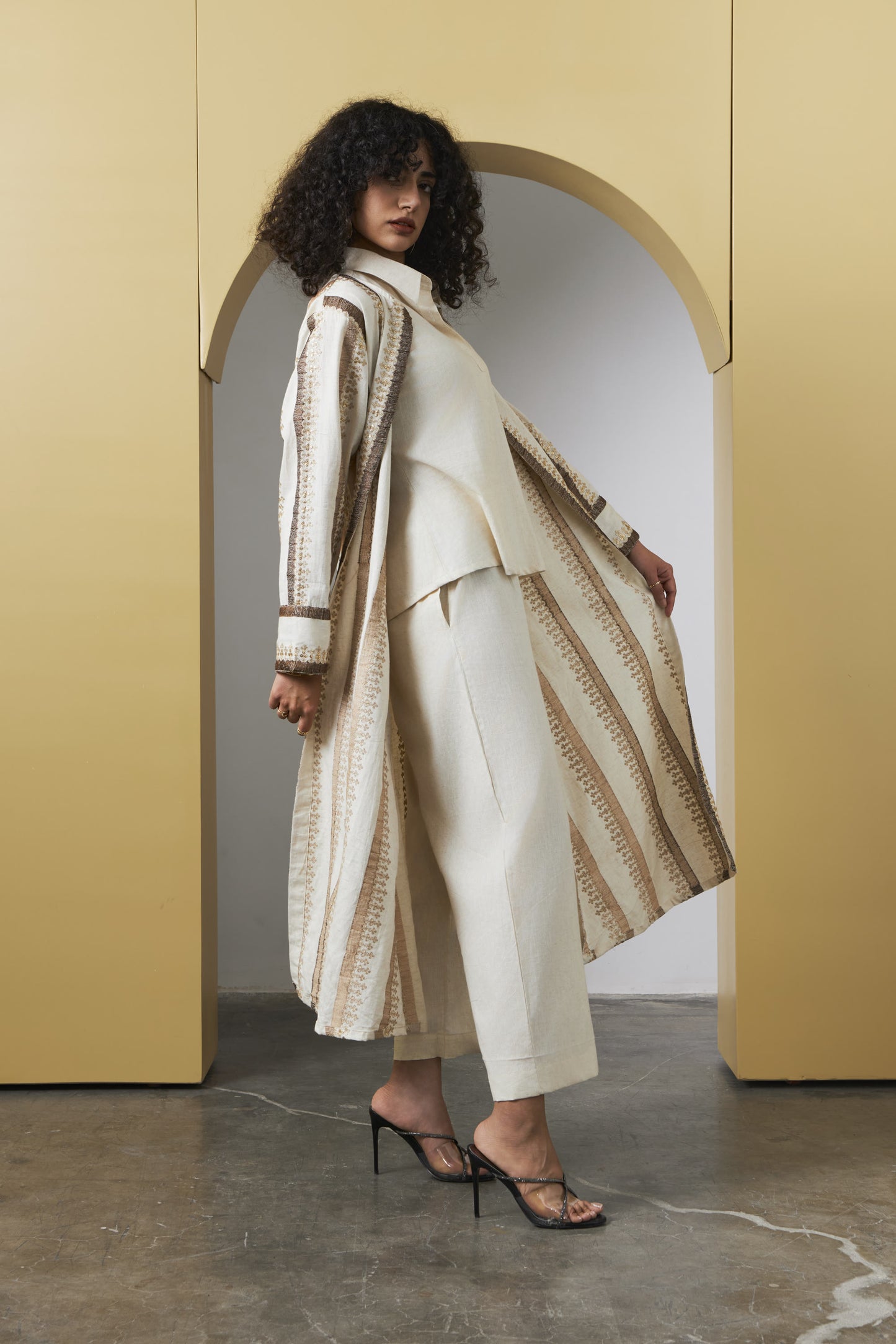 A model with curly dark hair stands in a minimalist studio setting with a yellow archway background, wearing a cream and beige patterned long cardigan over a matching cream top and cropped trousers, paired with black strappy heels.