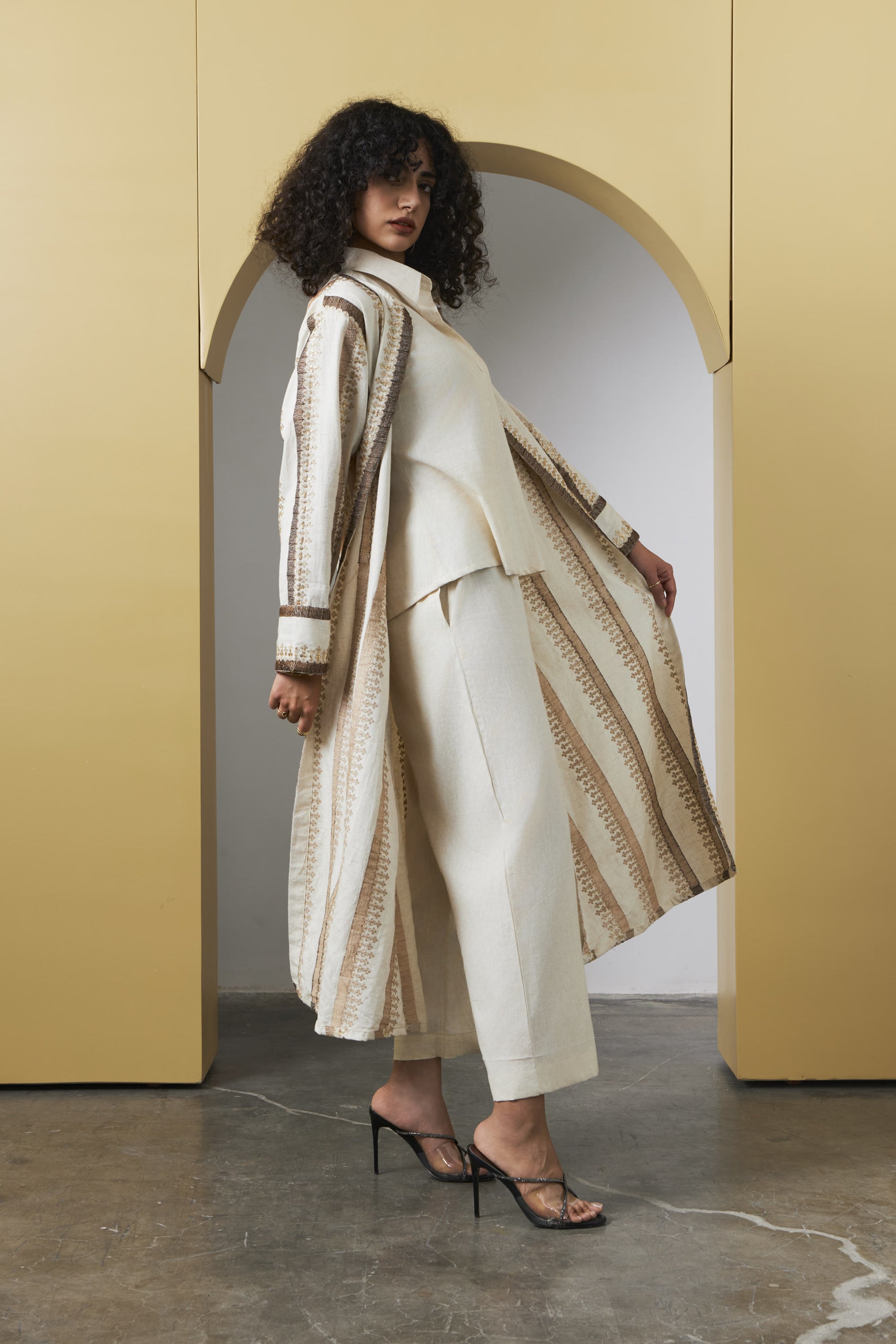 A model with curly dark hair stands in a minimalist studio setting with a yellow archway background, wearing a cream and beige patterned long cardigan over a matching cream top and cropped trousers, paired with black strappy heels.