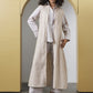A woman stands in a doorway wearing a long cream and beige vertically striped cardigan over a white shirt and matching wide-leg pants, paired with black heeled sandals.
