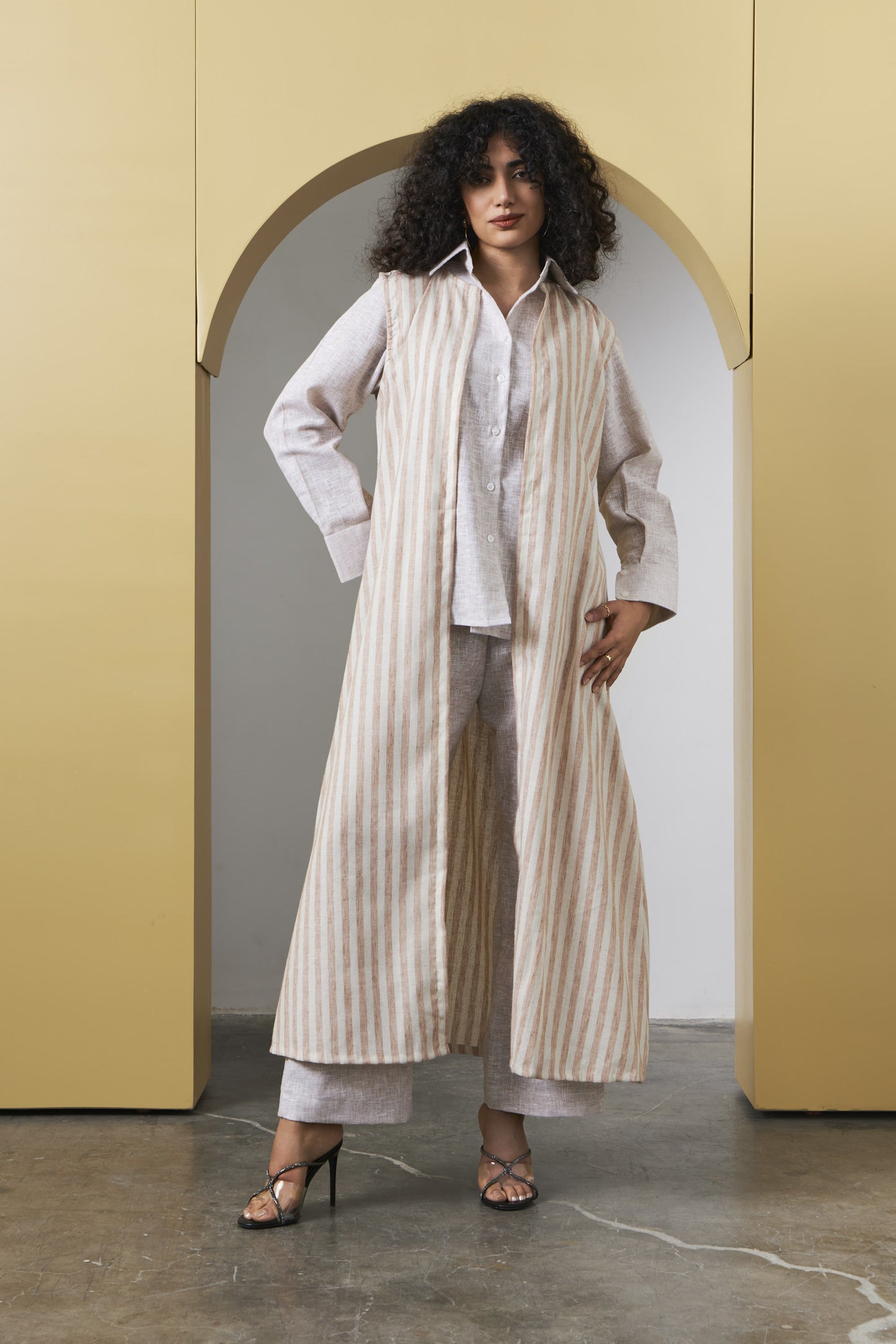 A woman stands in a doorway wearing a long cream and beige vertically striped cardigan over a white shirt and matching wide-leg pants, paired with black heeled sandals.
