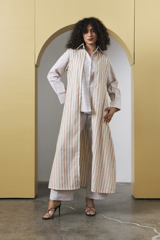 A woman stands in a doorway wearing a long cream and beige vertically striped cardigan over a white shirt and matching wide-leg pants, paired with black heeled sandals.