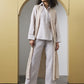 A woman models a light beige linen three-piece outfit consisting of a long-sleeve button-up shirt, matching wide-leg trousers, and a long open-front cardigan with vertical stripes, standing in front of a yellow arched wall.