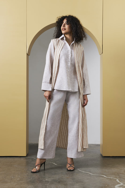 A woman models a light beige linen three-piece outfit consisting of a long-sleeve button-up shirt, matching wide-leg trousers, and a long open-front cardigan with vertical stripes, standing in front of a yellow arched wall.