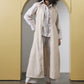 A woman stands in a studio setting with a yellow arched backdrop, wearing a long cream and beige vertically striped linen-blend vest over a white button-up shirt, light gray wide-leg pants, and black strappy heels.
