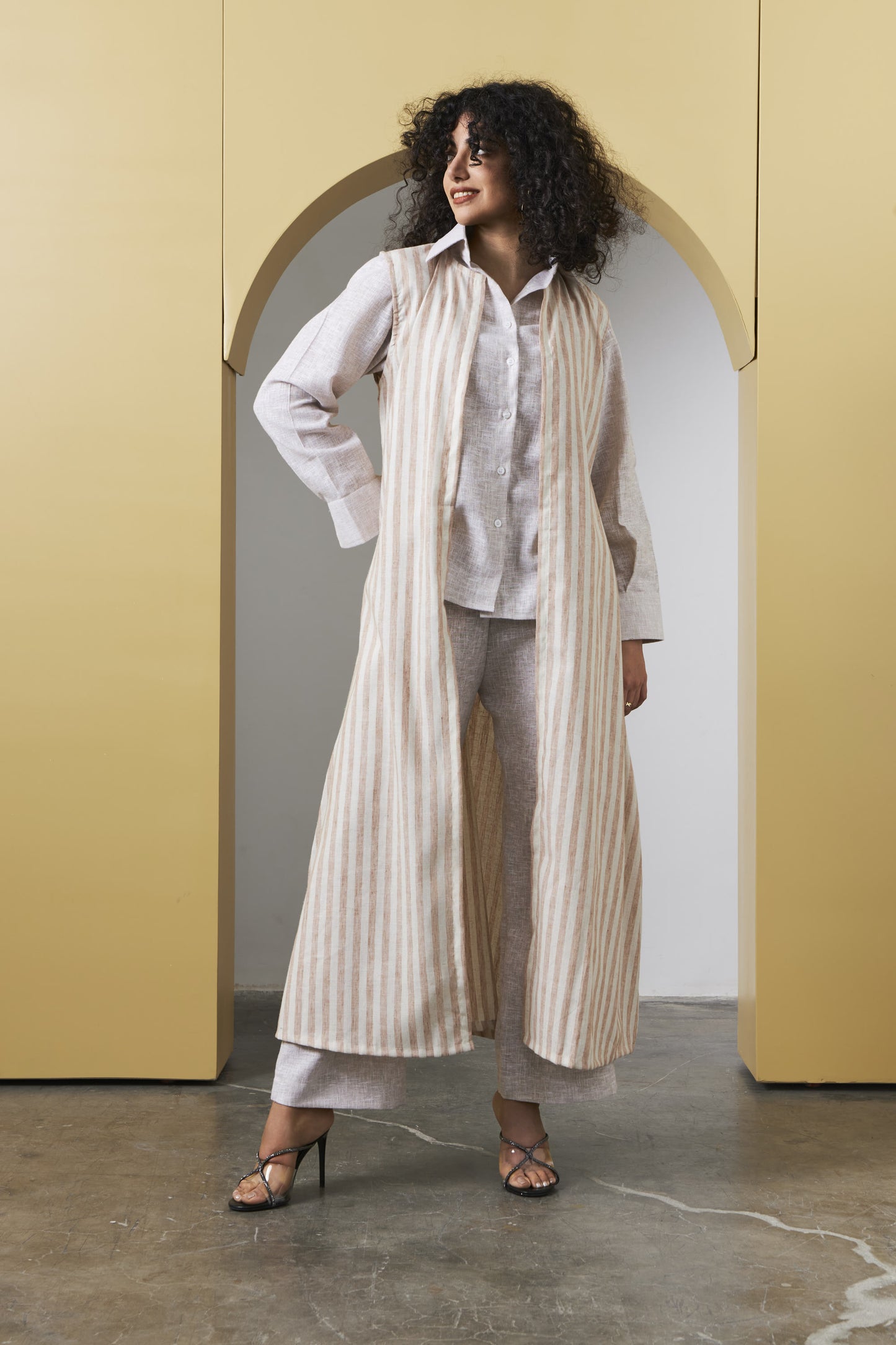 A woman stands in a studio setting with a yellow arched backdrop, wearing a long cream and beige vertically striped linen-blend vest over a white button-up shirt, light gray wide-leg pants, and black strappy heels.