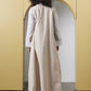 A woman viewed from behind wearing a long cream and beige vertically striped tunic with white sleeves, standing in front of a yellow architectural archway.