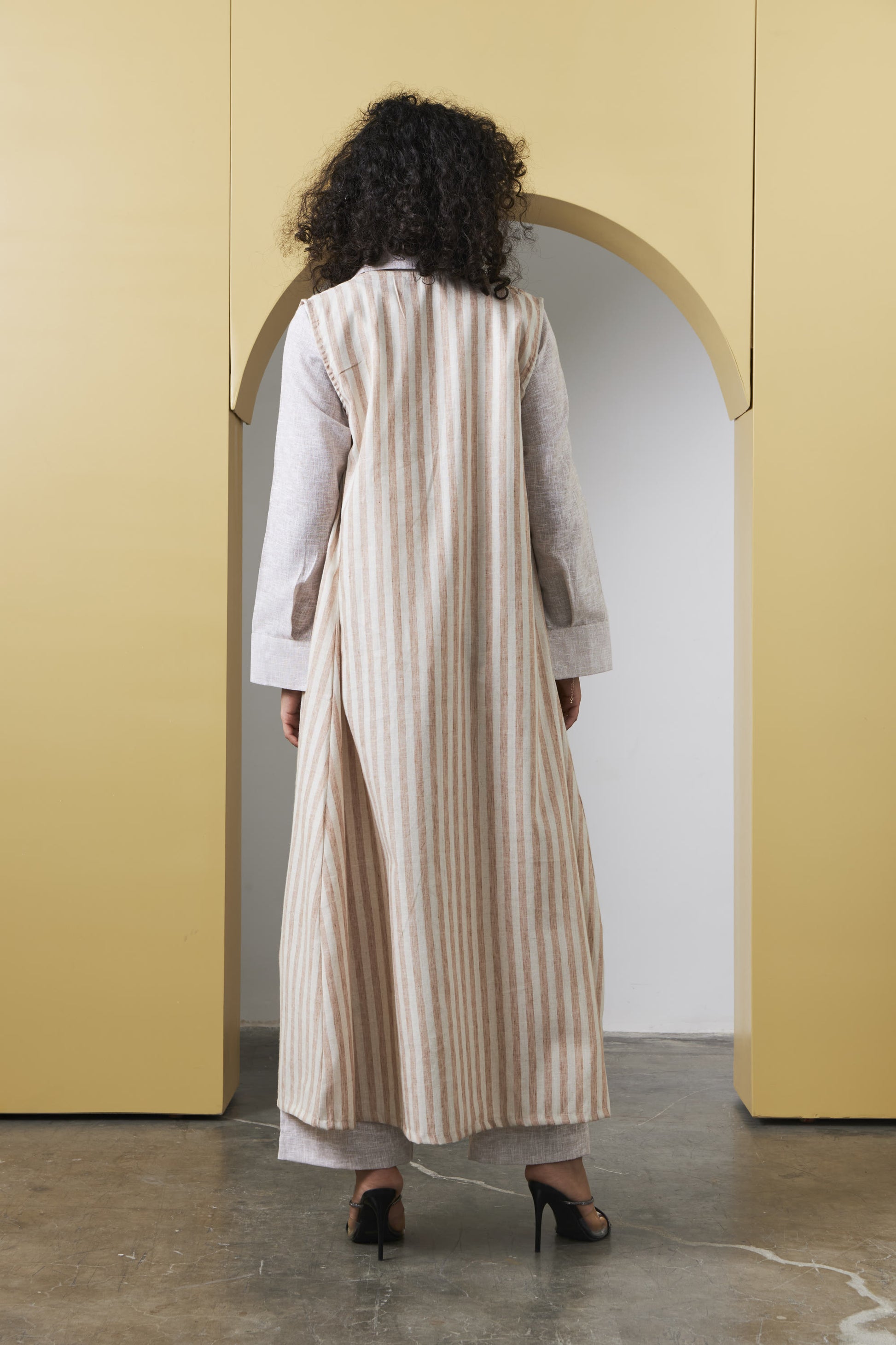 A woman viewed from behind wearing a long cream and beige vertically striped tunic with white sleeves, standing in front of a yellow architectural archway.