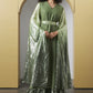 A woman stands in a studio wearing a flowing sage green dress with a wide metallic green cape and a matching waist belt, posed in front of a beige arched backdrop.