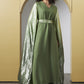 A woman wearing a long olive green dress with a shiny metallic green cape and a matching waist belt, standing in front of a beige arched backdrop.