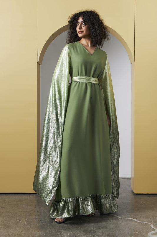 A woman wearing a long olive green dress with a shiny metallic green cape and a matching waist belt, standing in front of a beige arched backdrop.
