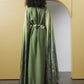 A model viewed from behind wearing a long olive green dress with a matching satin waist tie and a shimmering metallic green cape, standing in front of a minimalist beige wall with an archway.