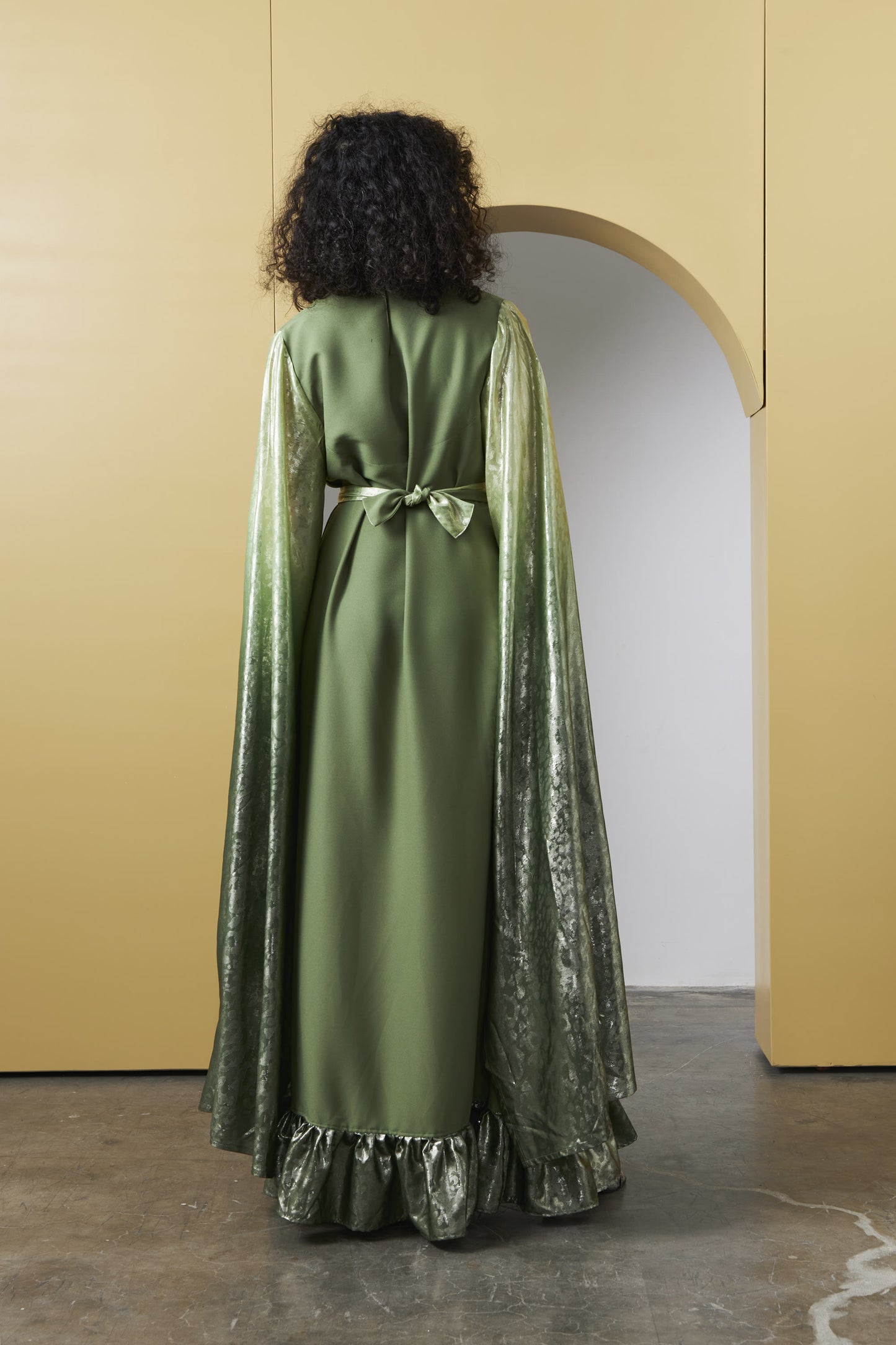 A model viewed from behind wearing a long olive green dress with a matching satin waist tie and a shimmering metallic green cape, standing in front of a minimalist beige wall with an archway.
