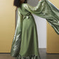 A woman models a sage green satin maxi dress with a matching metallic green brocade cape and a ruffled hem, set against a minimalist yellow and white background.