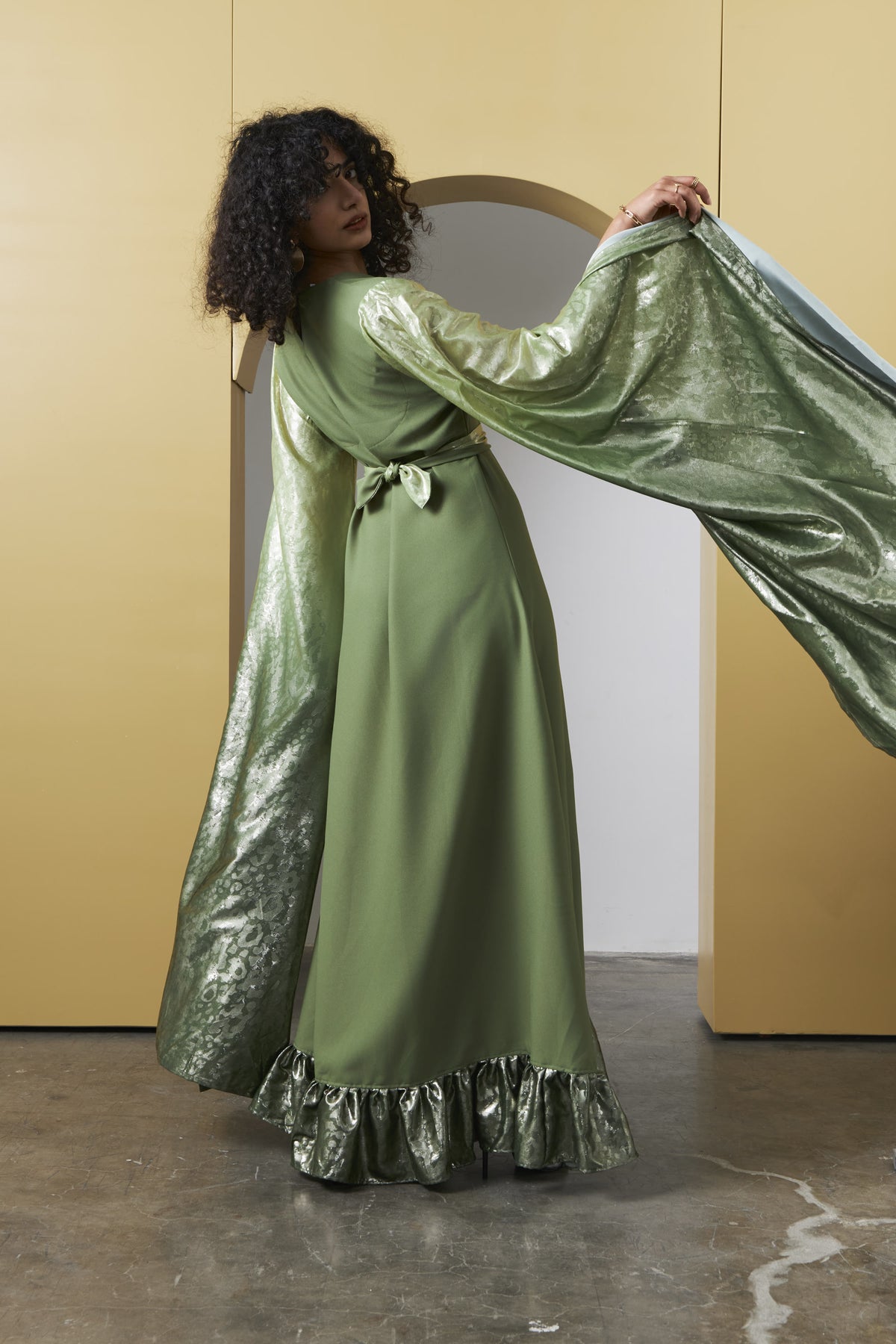 A woman models a sage green satin maxi dress with a matching metallic green brocade cape and a ruffled hem, set against a minimalist yellow and white background.
