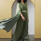 A woman modeling a flowing olive green maxi dress with wide draped sleeves, a matching belt, and a metallic green ruffled hem, posed in front of a yellow architectural archway.