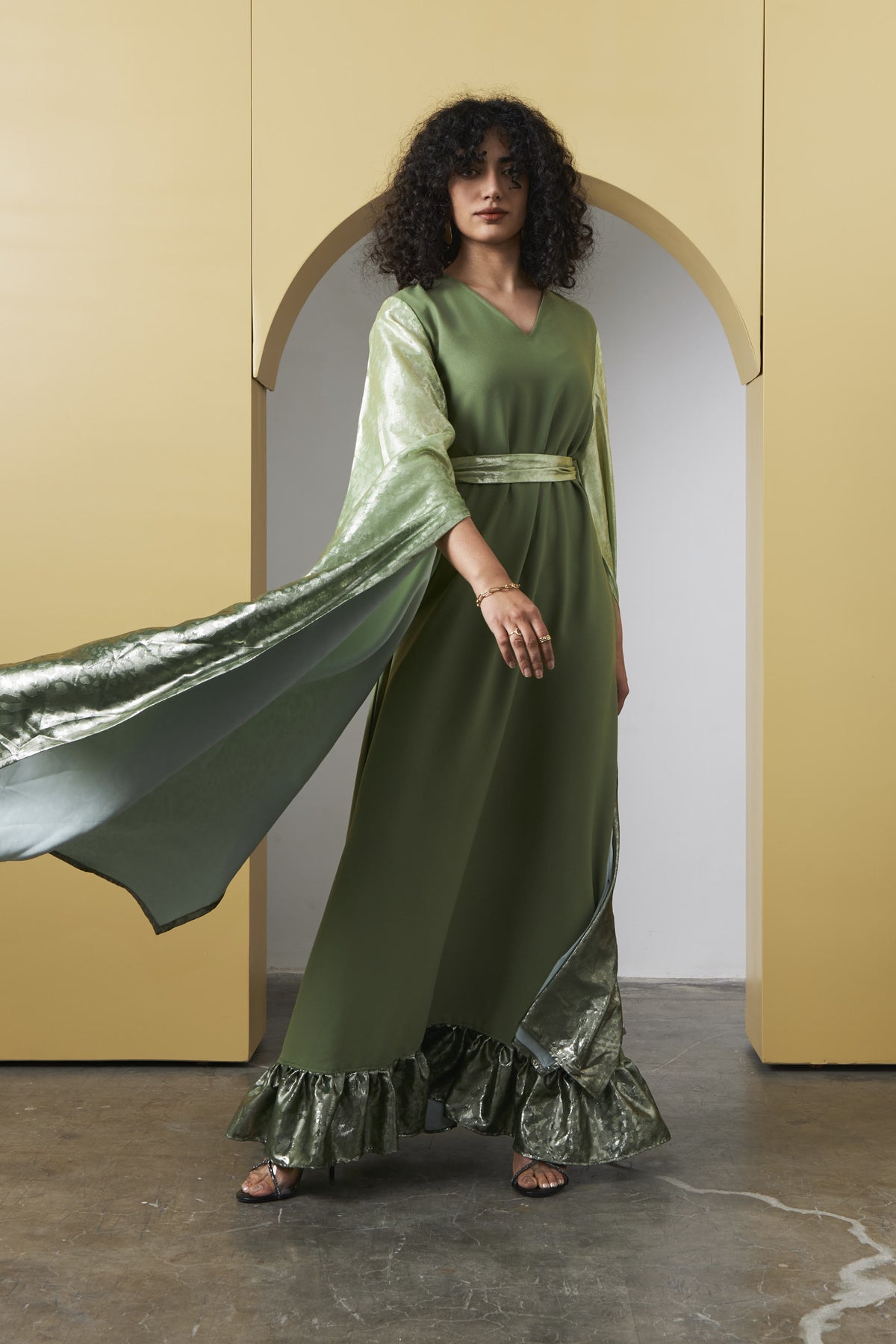 A woman modeling a flowing olive green maxi dress with wide draped sleeves, a matching belt, and a metallic green ruffled hem, posed in front of a yellow architectural archway.