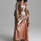 A model with curly black hair wearing a long, draped metallic rose gold gown with wide sleeves, standing against a neutral gray studio background.