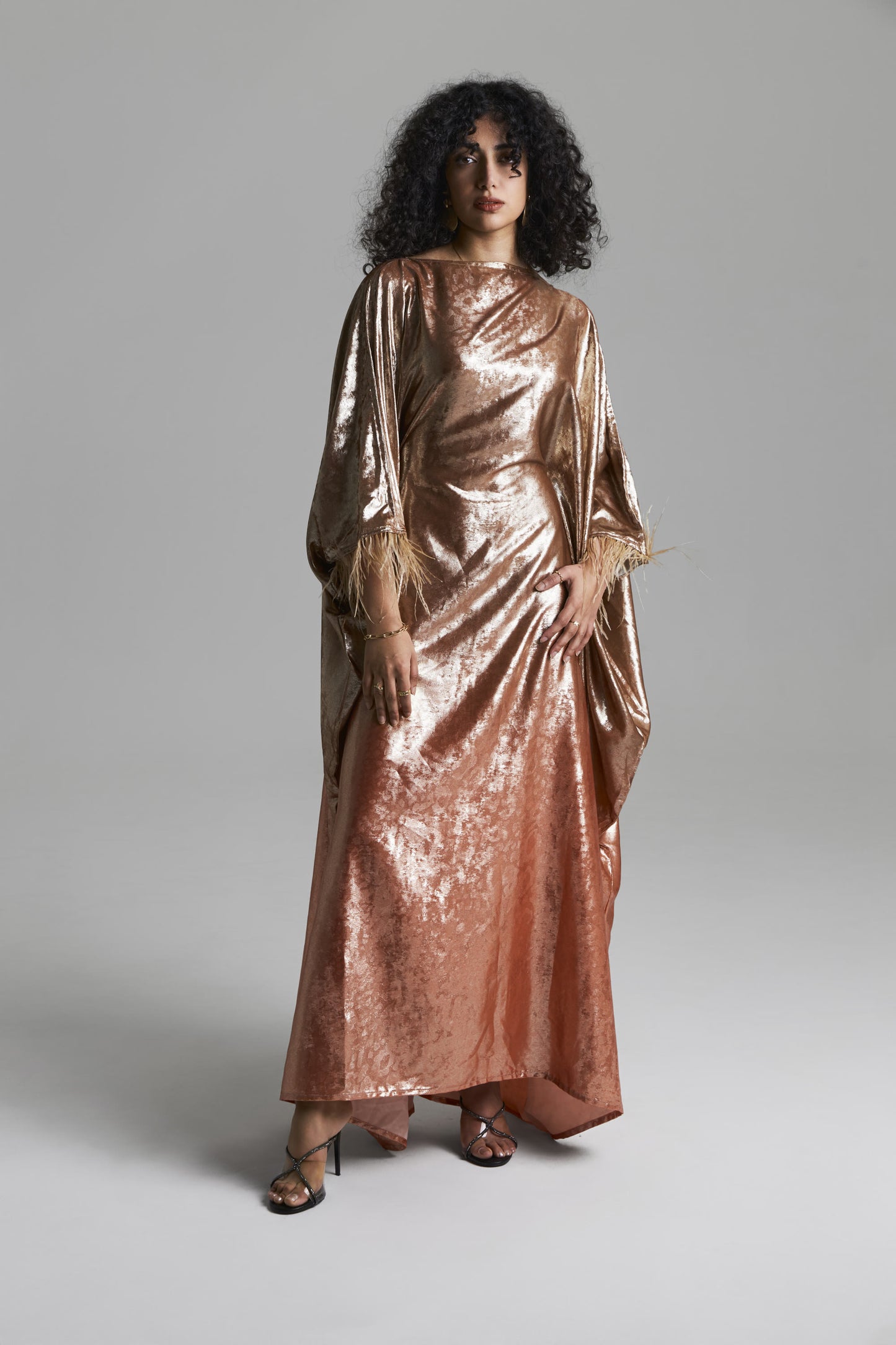 A model with curly black hair wearing a long, draped metallic rose gold gown with wide sleeves, standing against a neutral gray studio background.