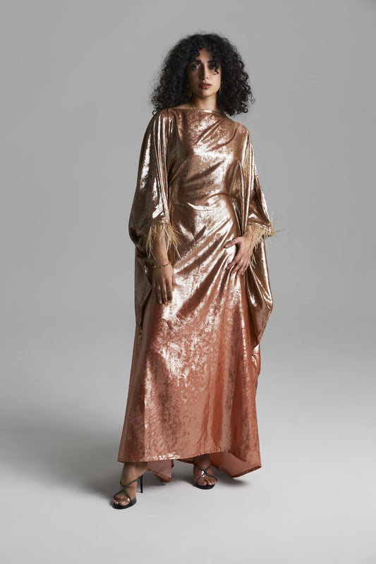 A model with curly black hair wearing a long, draped metallic rose gold gown with wide sleeves, standing against a neutral gray studio background.