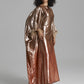 A model wearing a floor-length, metallic rose gold gown with wide draped sleeves and feathered cuffs, standing against a neutral gray studio background.