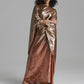 A model with curly black hair wearing a floor-length, metallic bronze satin gown with draped cape-like sleeves, standing against a neutral gray studio background.