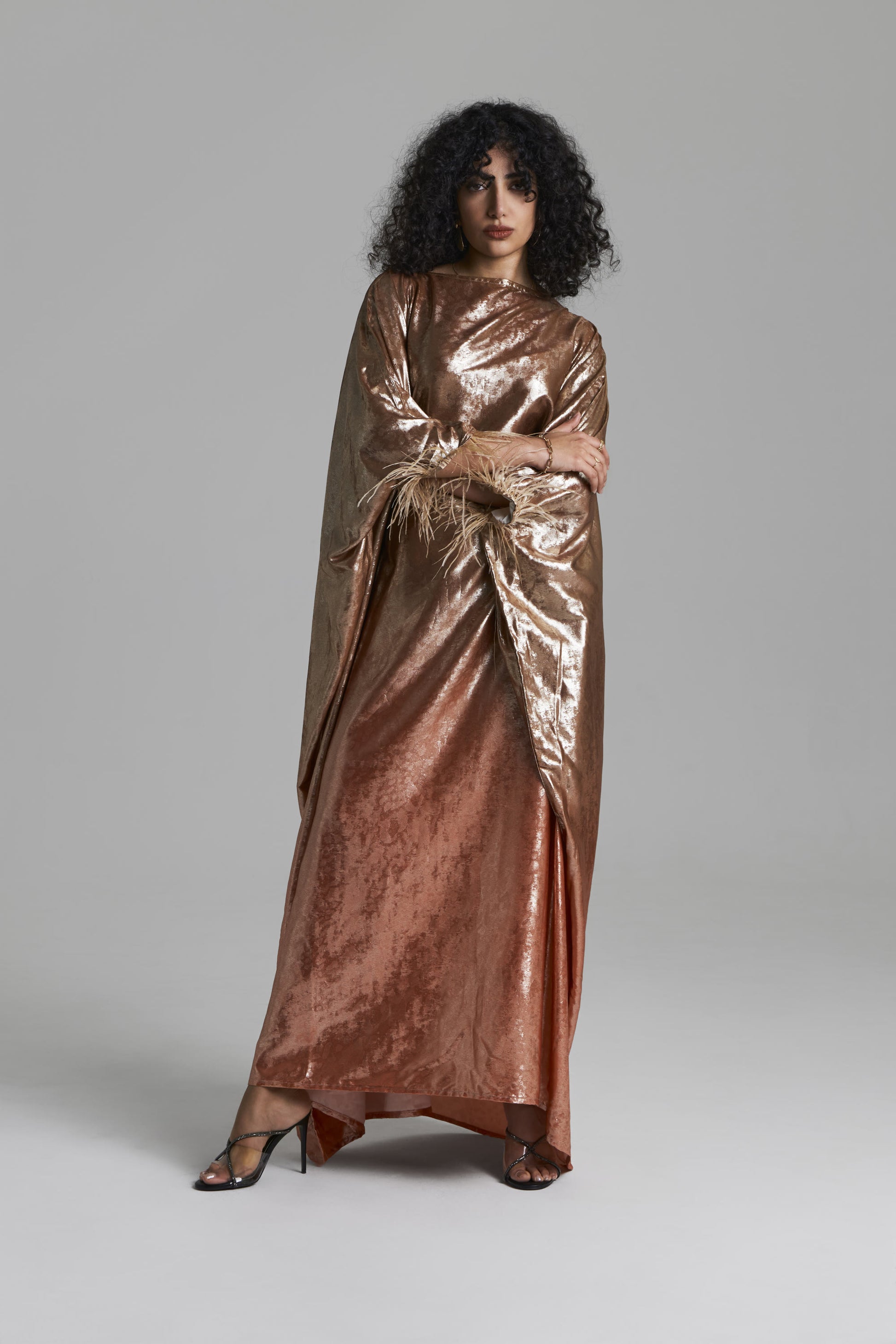 A model with curly black hair wearing a floor-length, metallic bronze satin gown with draped cape-like sleeves, standing against a neutral gray studio background.