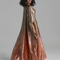 A woman modeling a floor-length metallic gown with a draped cape-like overlay, shown against a plain light gray studio background.