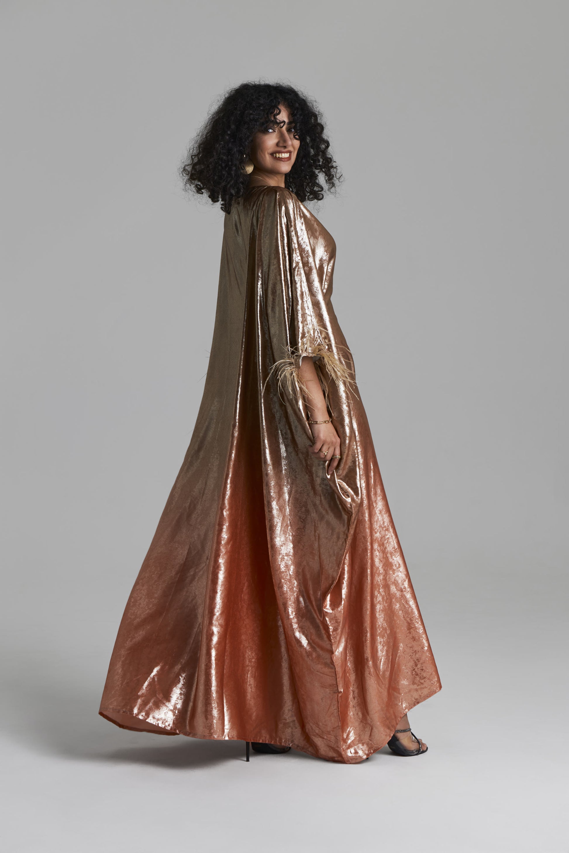 A woman modeling a floor-length metallic gown with a draped cape-like overlay, shown against a plain light gray studio background.