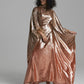 A model wearing a floor-length, metallic rose gold caftan gown with wide draped sleeves, standing against a neutral gray studio background.