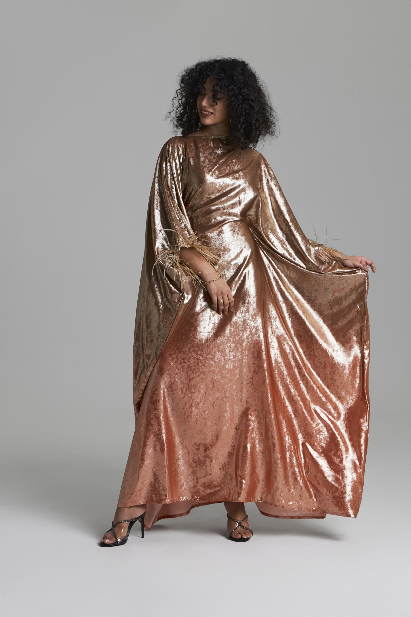 A model wearing a floor-length, metallic rose gold caftan gown with wide draped sleeves, standing against a neutral gray studio background.