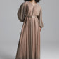 A model with curly black hair wearing a long-sleeved, floor-length taupe maxi dress with a matching belt, photographed against a neutral gray studio background.