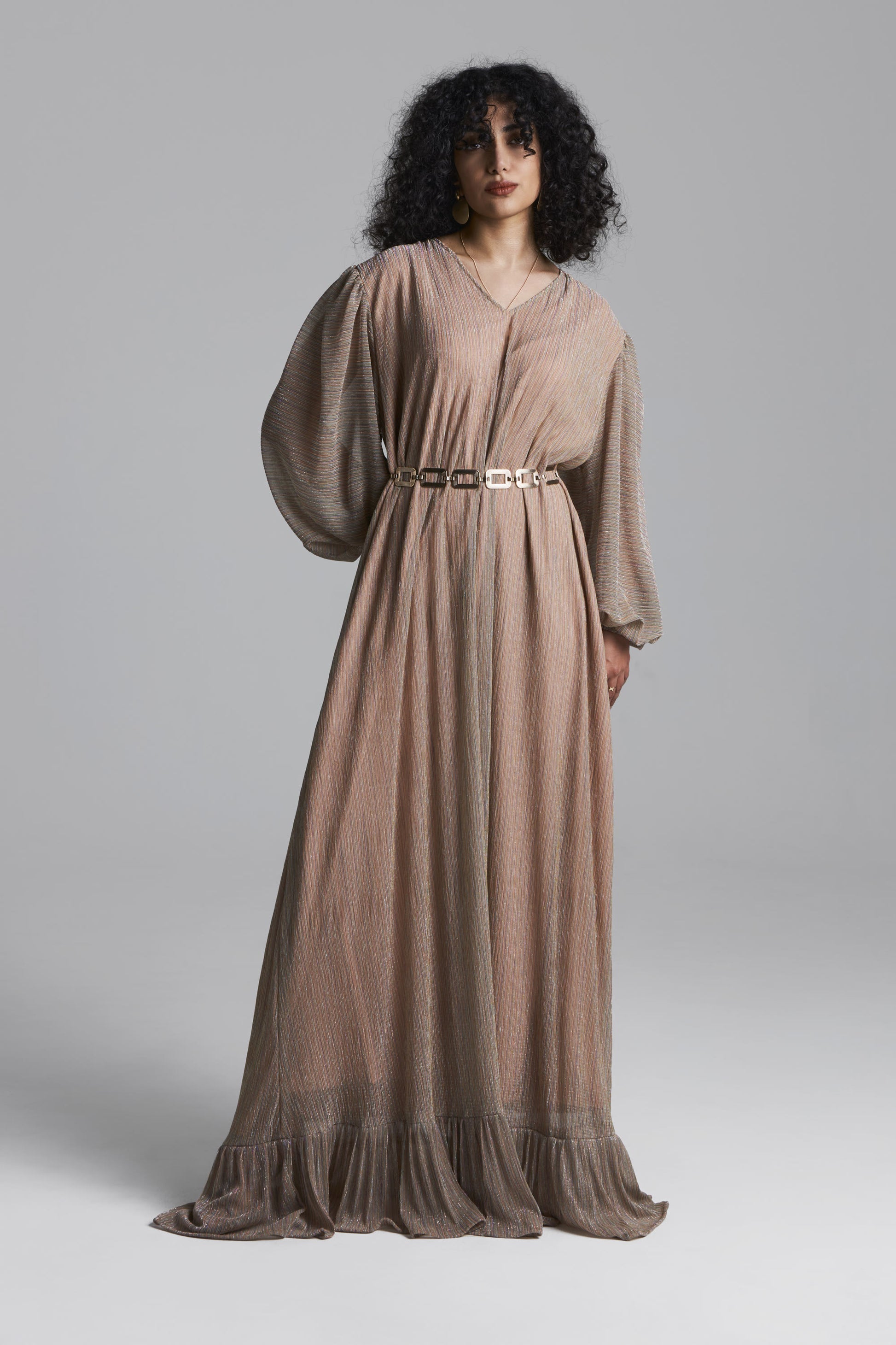A model with curly black hair wearing a long-sleeved, floor-length taupe maxi dress with a matching belt, photographed against a neutral gray studio background.