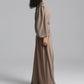A model with curly black hair wearing a long-sleeved, floor-length taupe maxi dress with a matching belt, side view photographed against a neutral gray studio background.
